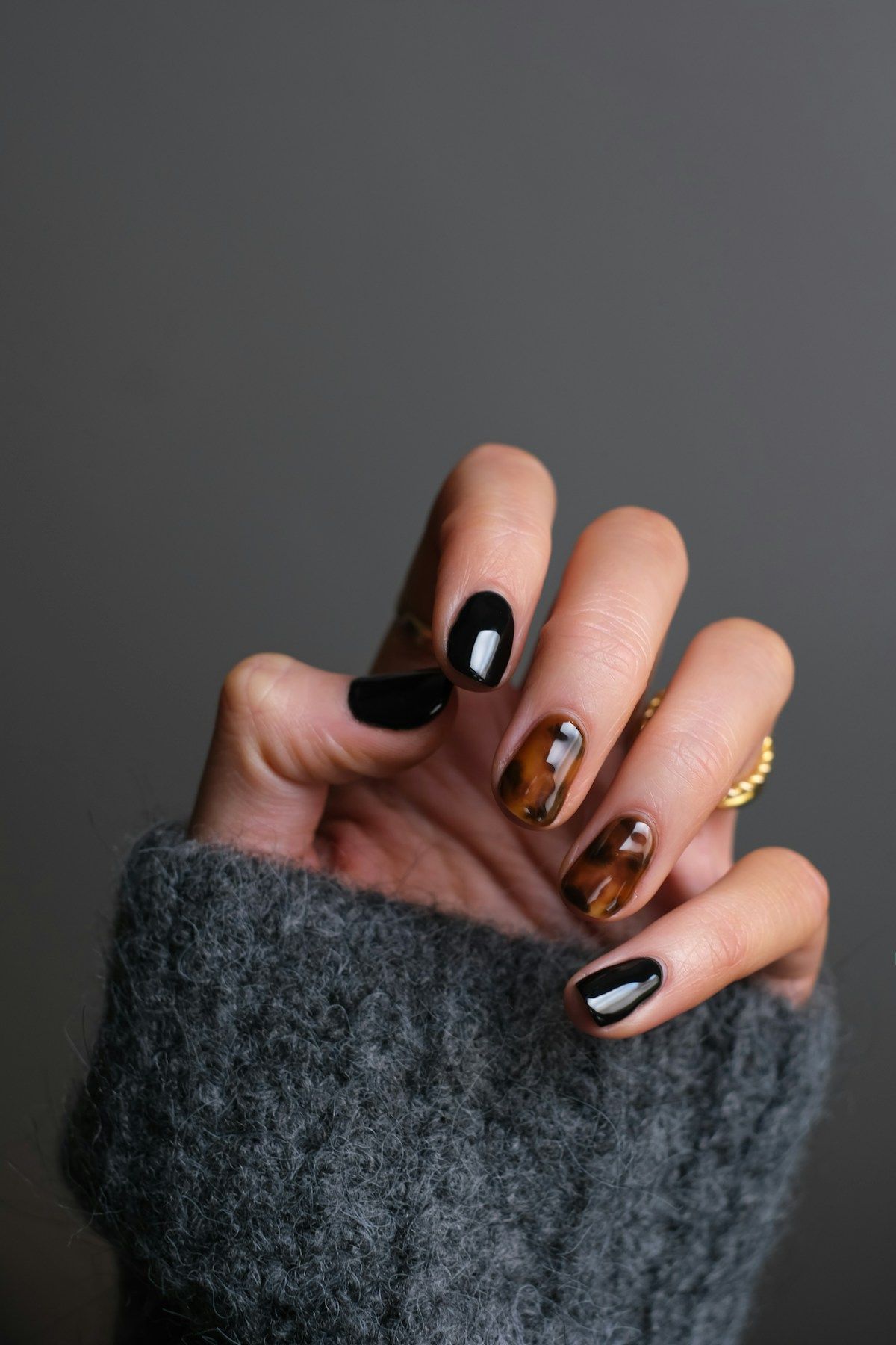 Mano con manicura en tonos negro y carey, con un anillo dorado, sobre un suéter gris