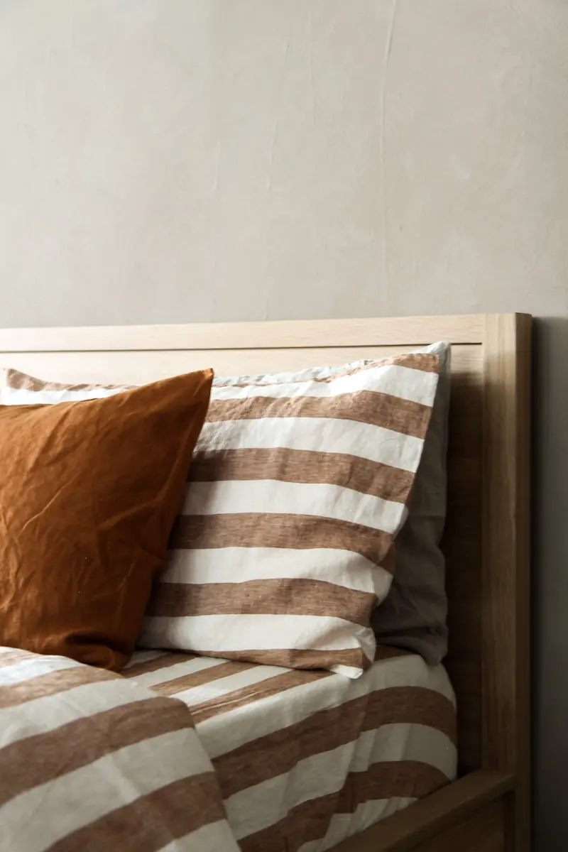 Detalle de ropa de cama a rayas con cojín color terracota sobre cabecera de madera